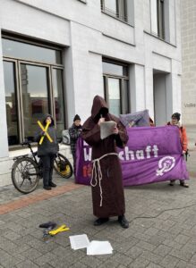 Mönch von Lützerath steht mit Mikro lesend vor Banner mit Aufschrift "Diese Wirtschaft tötet"